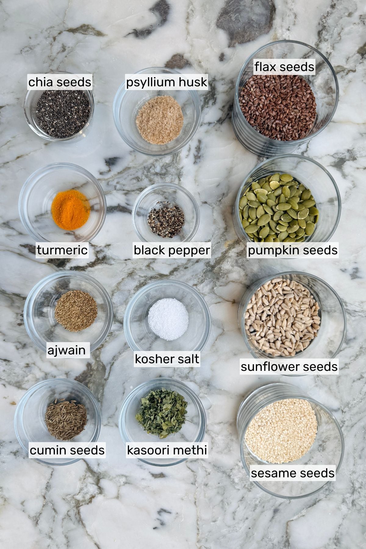 Twelve small glass bowls on a marble surface, each containing a labeled ingredient: chia seeds, psyllium husk, flax seeds, turmeric, black pepper, pumpkin seeds, ajwain, kosher salt, sunflower seeds, cumin seeds, kasoori methi, and sesame seeds.
