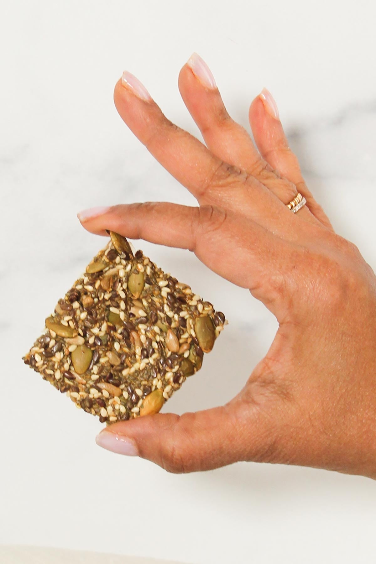 A hand holding a square, multigrain seed cracker against a light background.