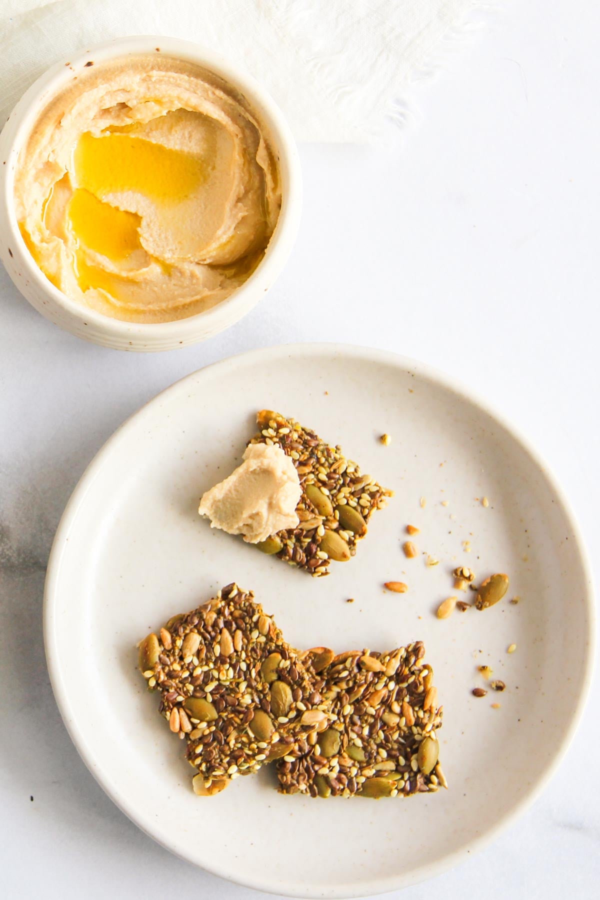 A bowl of hummus with olive oil sits next to a plate holding three seed crackers, one topped with a dollop of hummus.