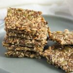 A stack of seed crackers sits on a gray plate, with additional crackers lying flat beside the stack.