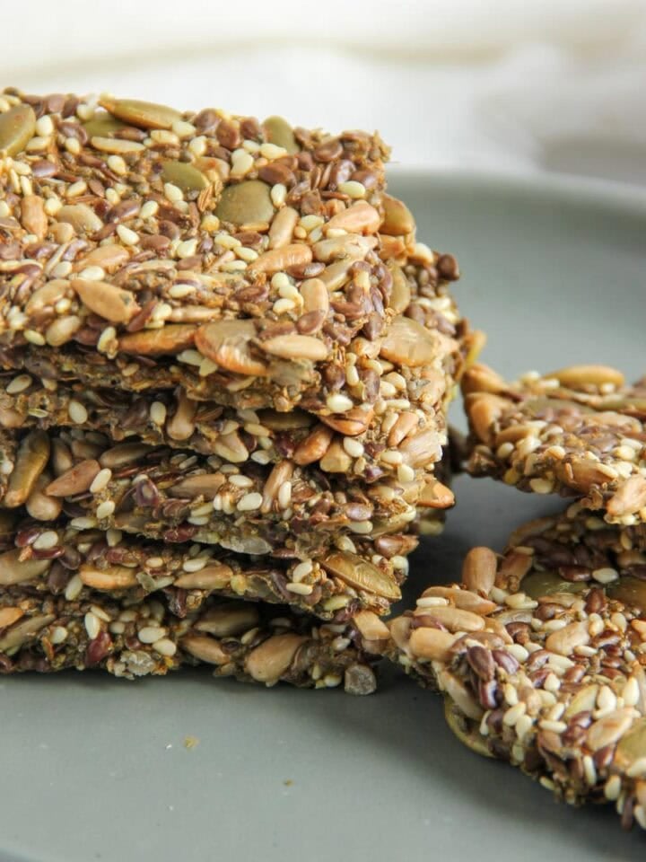 A stack of seed crackers sits on a gray plate, with additional crackers lying flat beside the stack.