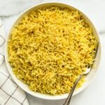 A white bowl filled with cooked yellow rice and quinoa, with a silver fork resting inside, placed on a white surface next to a white and gray checkered cloth.