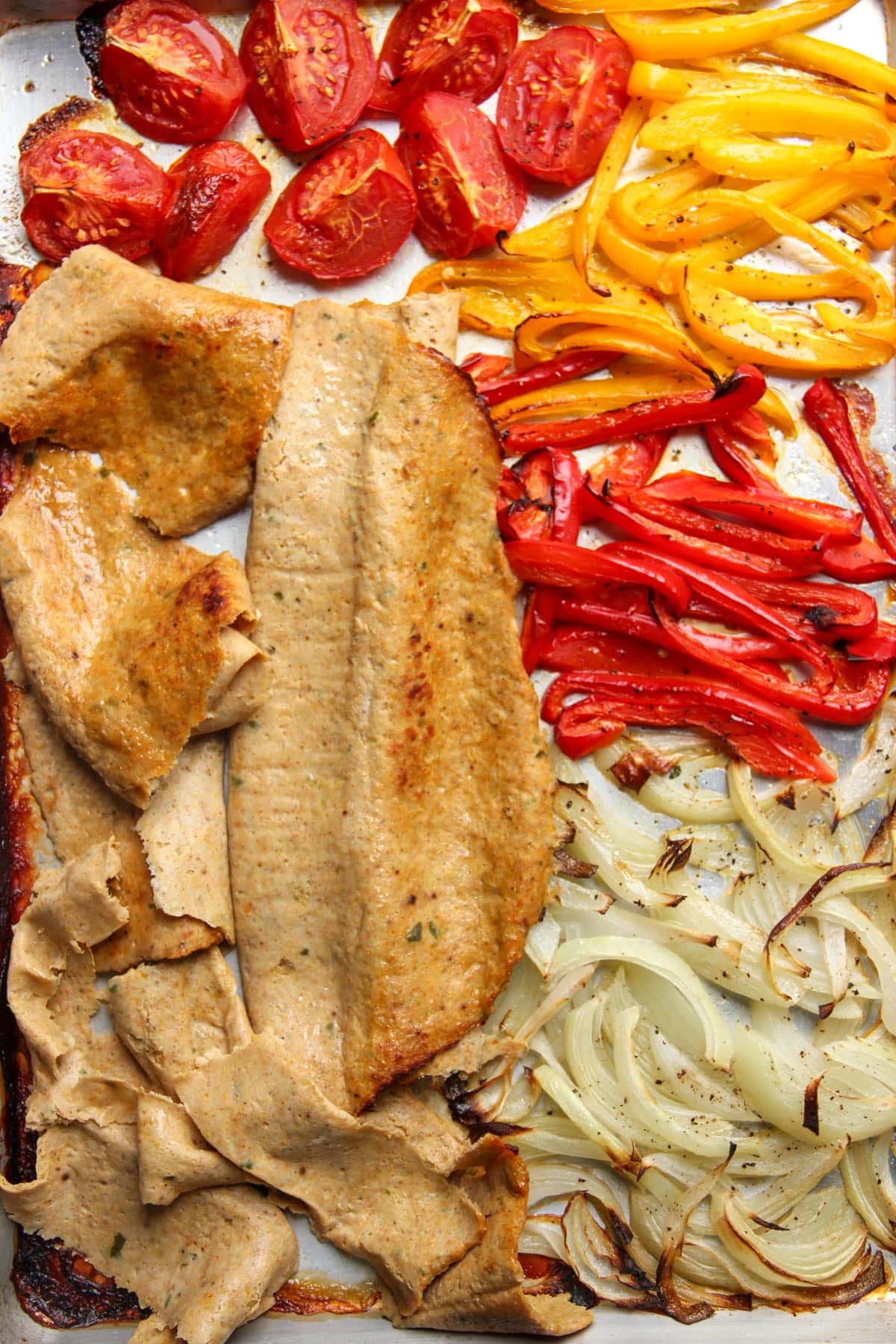 Seitán en rodajas, tomates asados, pimientos amarillos y rojos y cebollas dispuestos en una bandeja para horno.