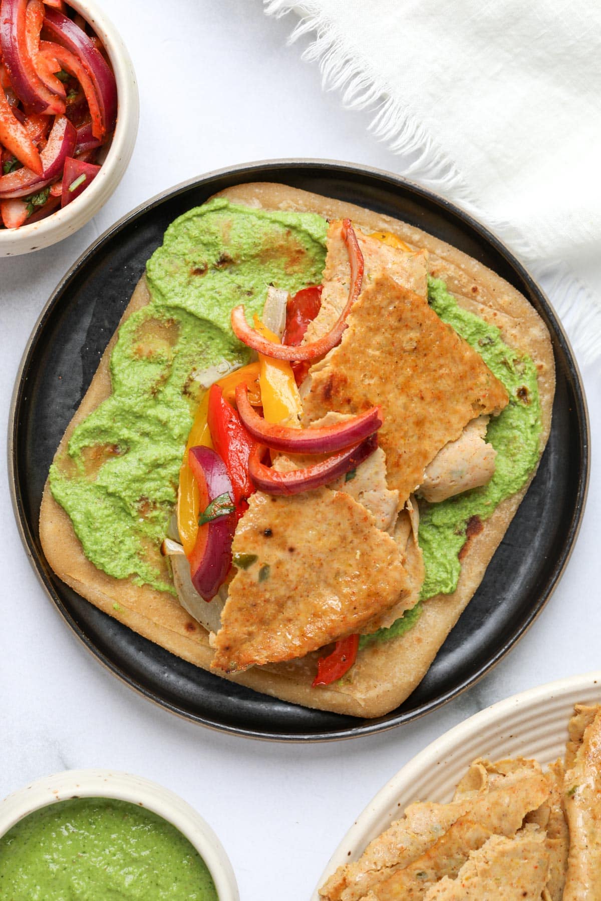 A square flatbread with pesto sauce, sliced ​​cooked meat, and colorful sautéed peppers and onions served on a black plate. There are small bowls nearby with extra sauce and vegetables.