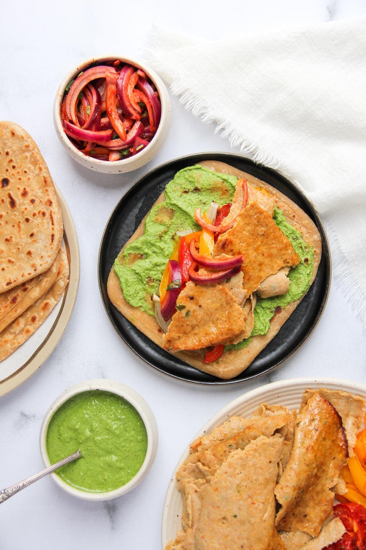 Un pan plano cubierto con salsa verde, seitán cocido en rodajas y pimientos mixtos, rodeado de panes planos adicionales, salsa y un plato de cebollas y pimientos en rodajas.