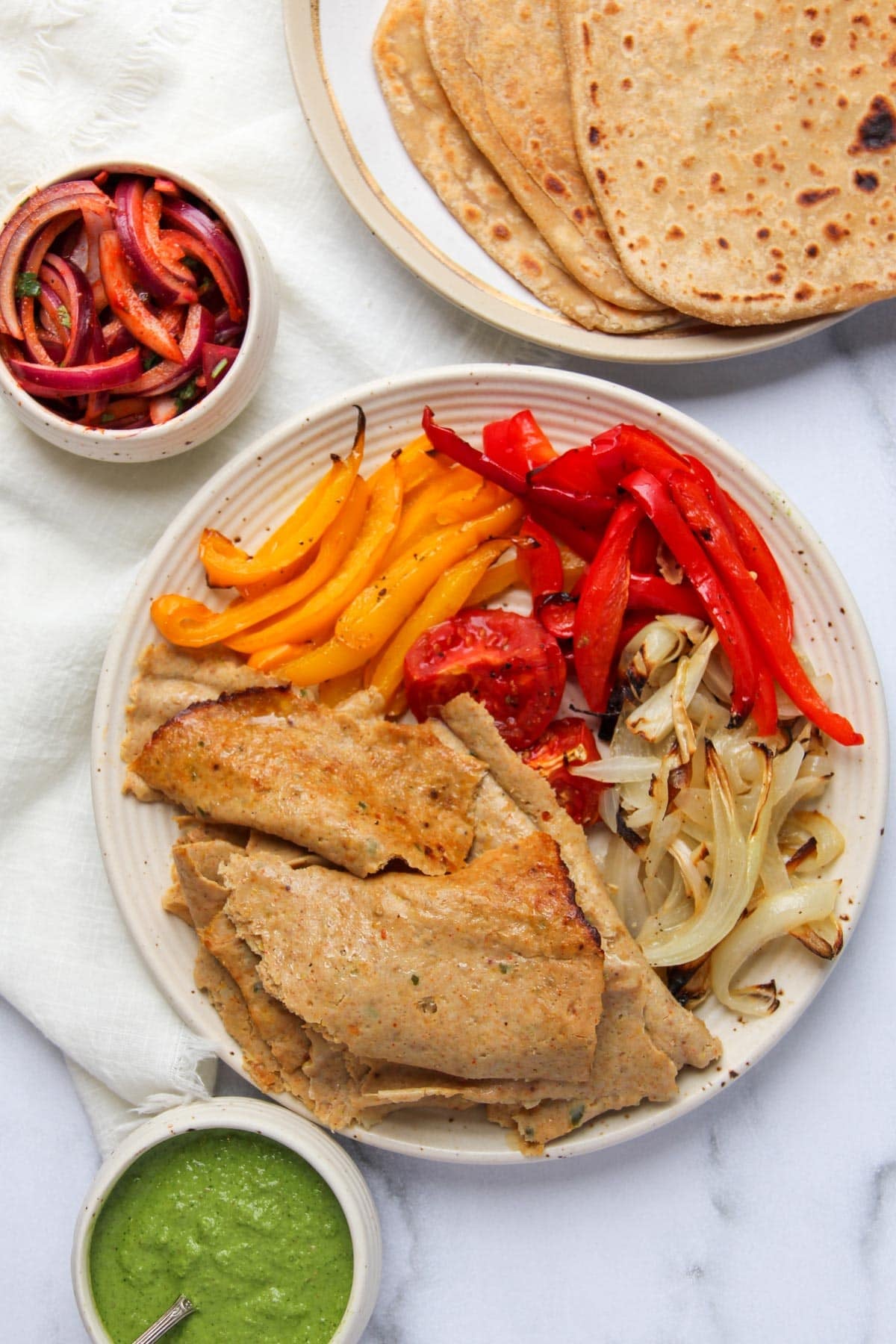 Un plato con rodajas de seitán, pimientos asados, cebolla y tomate, servido con panes planos, salsa verde y un plato de cebollas encurtidas sobre una superficie blanca.