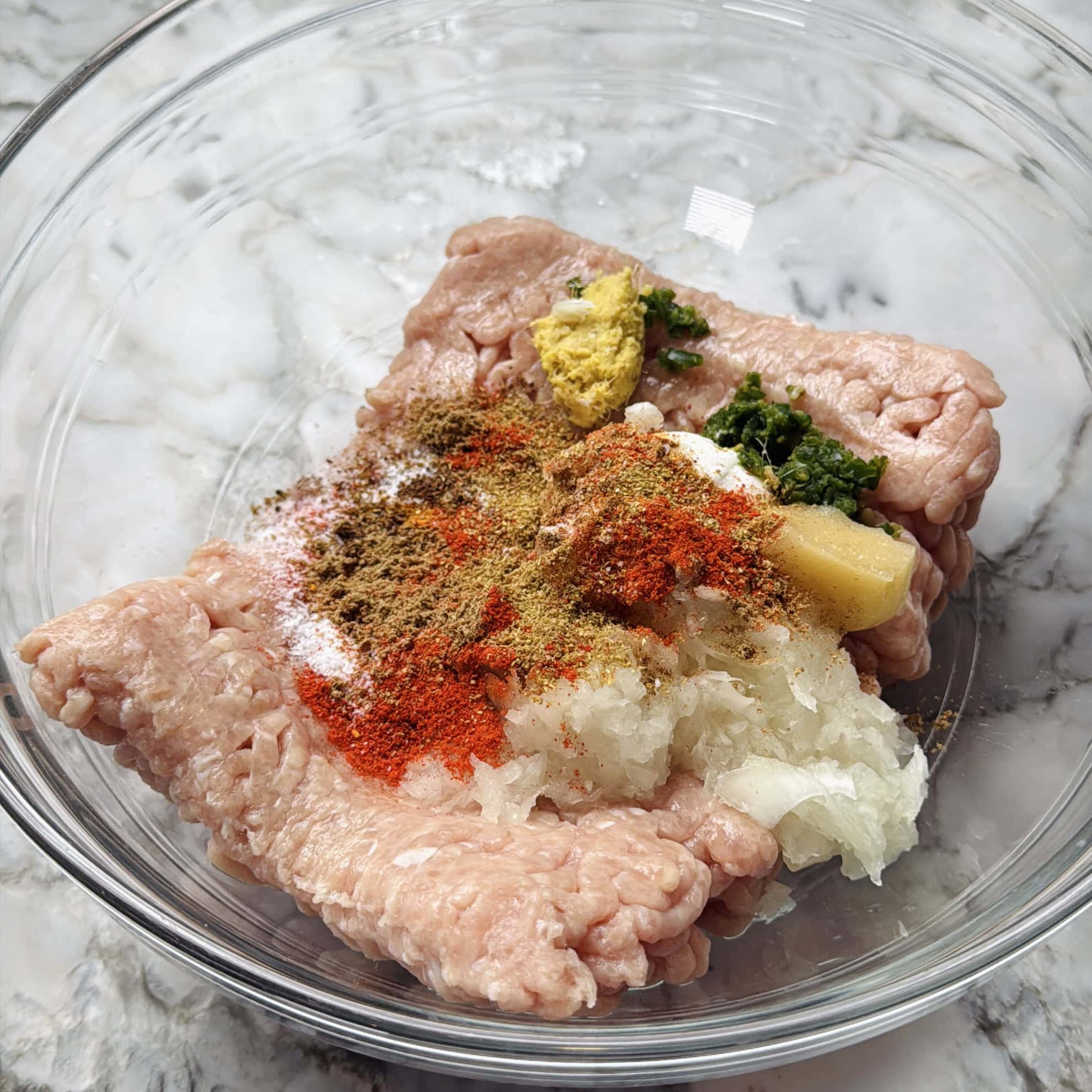 Carne picada cruda en un recipiente de vidrio adornada con cebolla rallada, hierbas verdes picadas, jengibre y diversas especias molidas sobre una encimera de mármol.