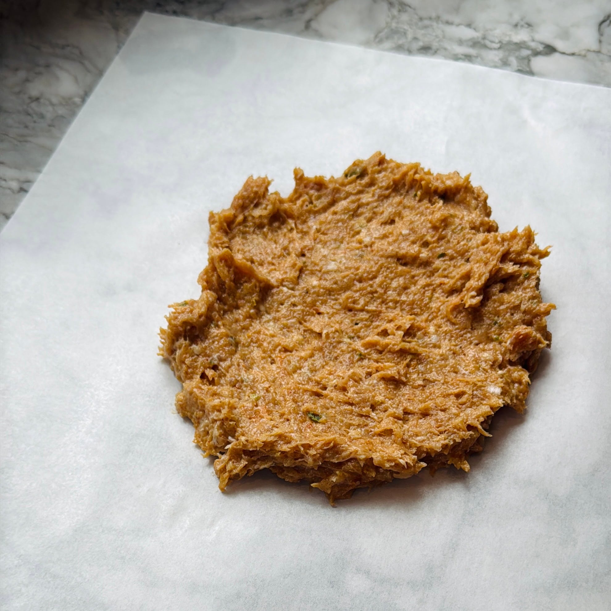 A round, flattened portion of brown, textured dough sits on a sheet of parchment paper on a marble surface.