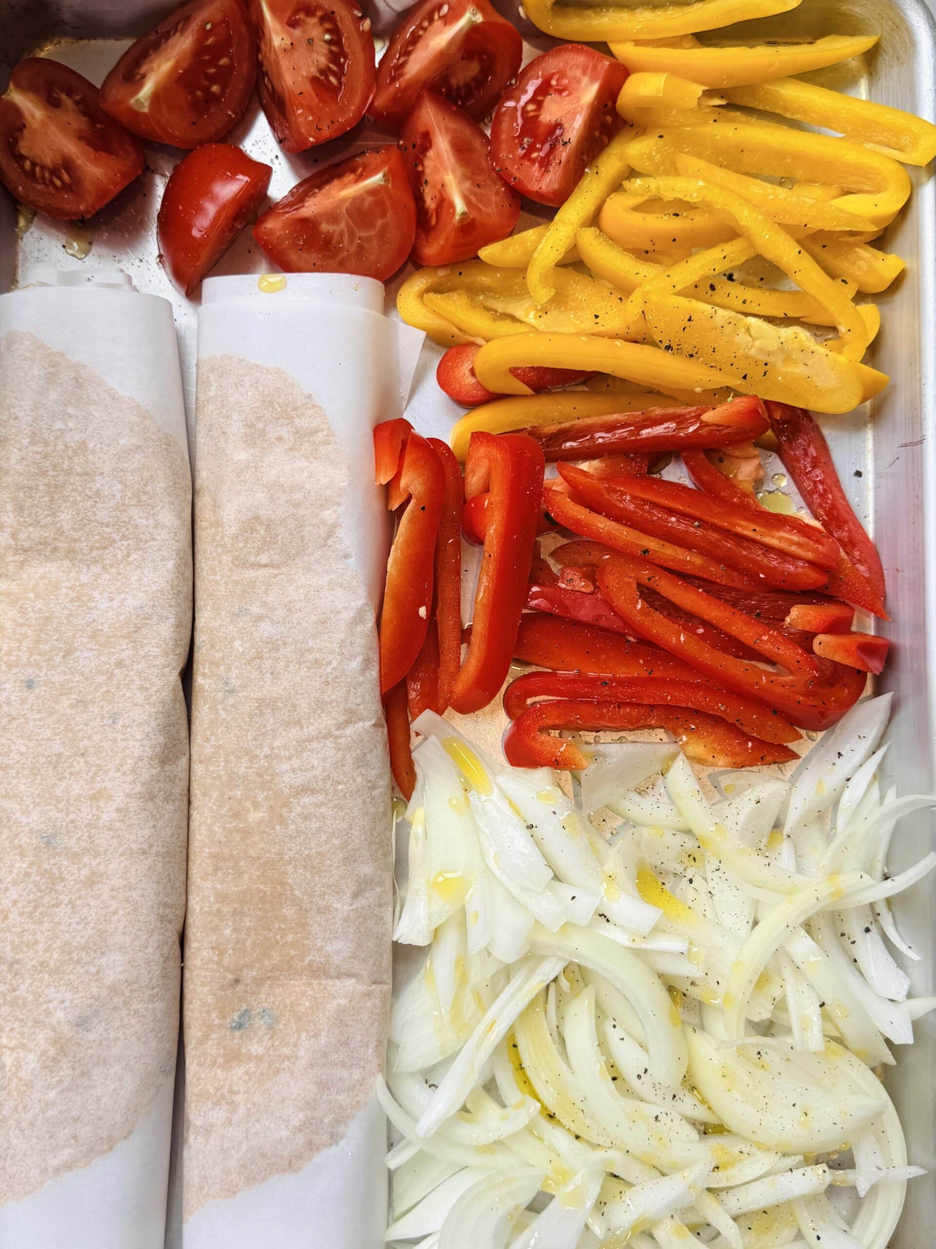 A tray with sliced tomatoes, yellow peppers, red peppers, onions, and two parchment-wrapped items, all seasoned with oil and pepper.