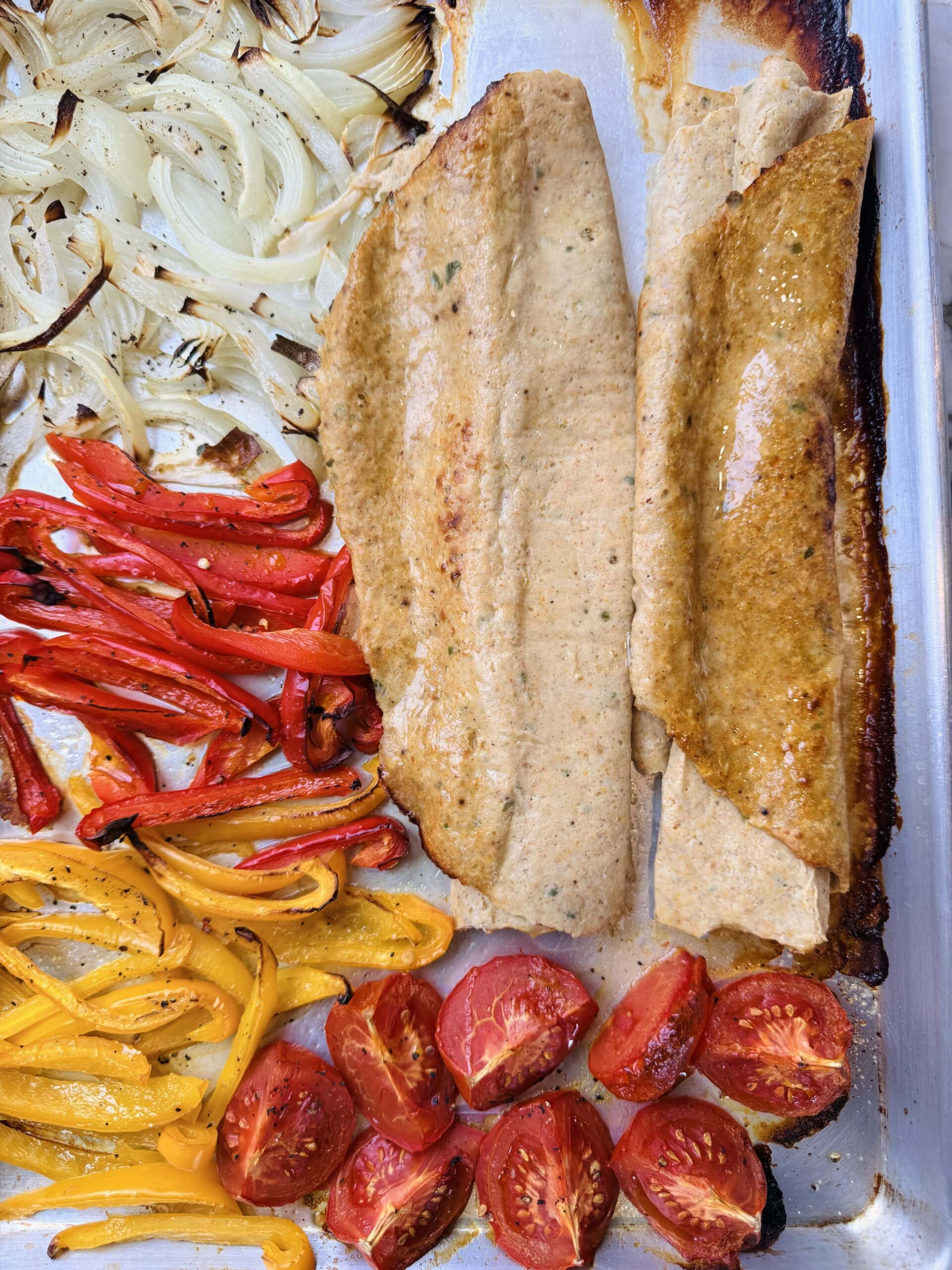 Baked white fish fillets on a sheet pan with roasted onions, red and yellow bell peppers, and halved tomatoes.