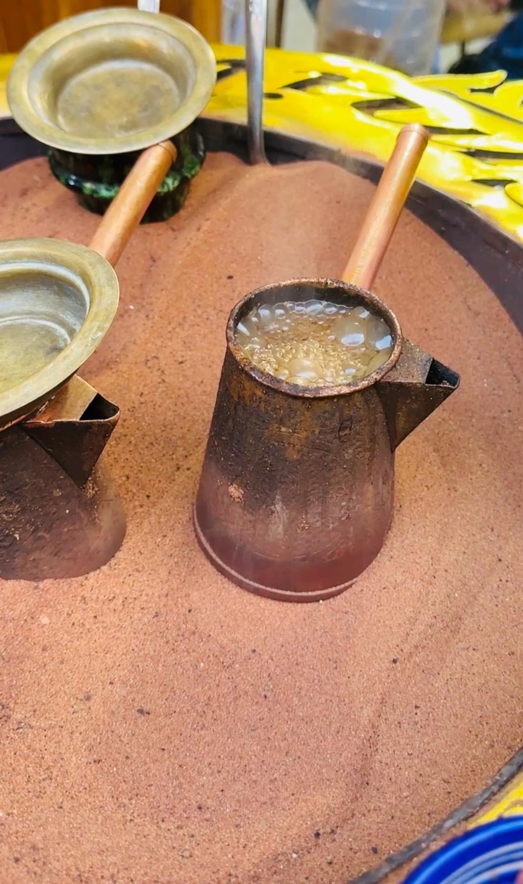 A metal pot with a wooden handle is brewing coffee in hot sand, next to a lid and another tool, illustrating a traditional sand coffee-making method.