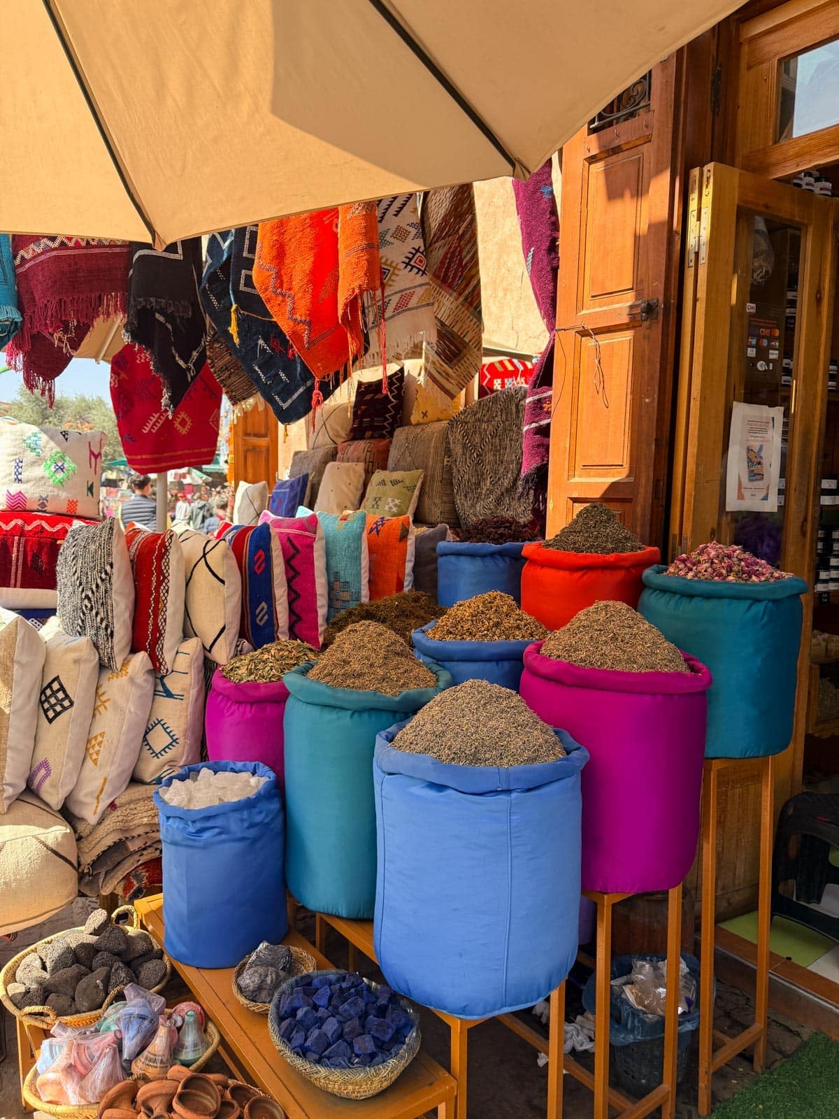 Colorful fabric bags filled with spices are displayed outside a shop selling woven rugs, pillows, and textiles, with more baskets and cushions visible inside.