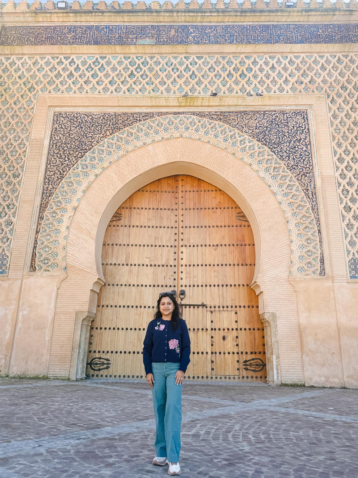 A person stands in front of a large ornate wooden gate set in an arched entrance with intricate blue and beige tilework.