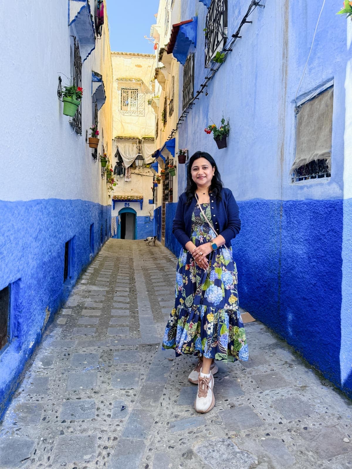 A woman in a floral dress stands smiling in a narrow alleyway with blue and white painted walls and potted plants on window ledges.