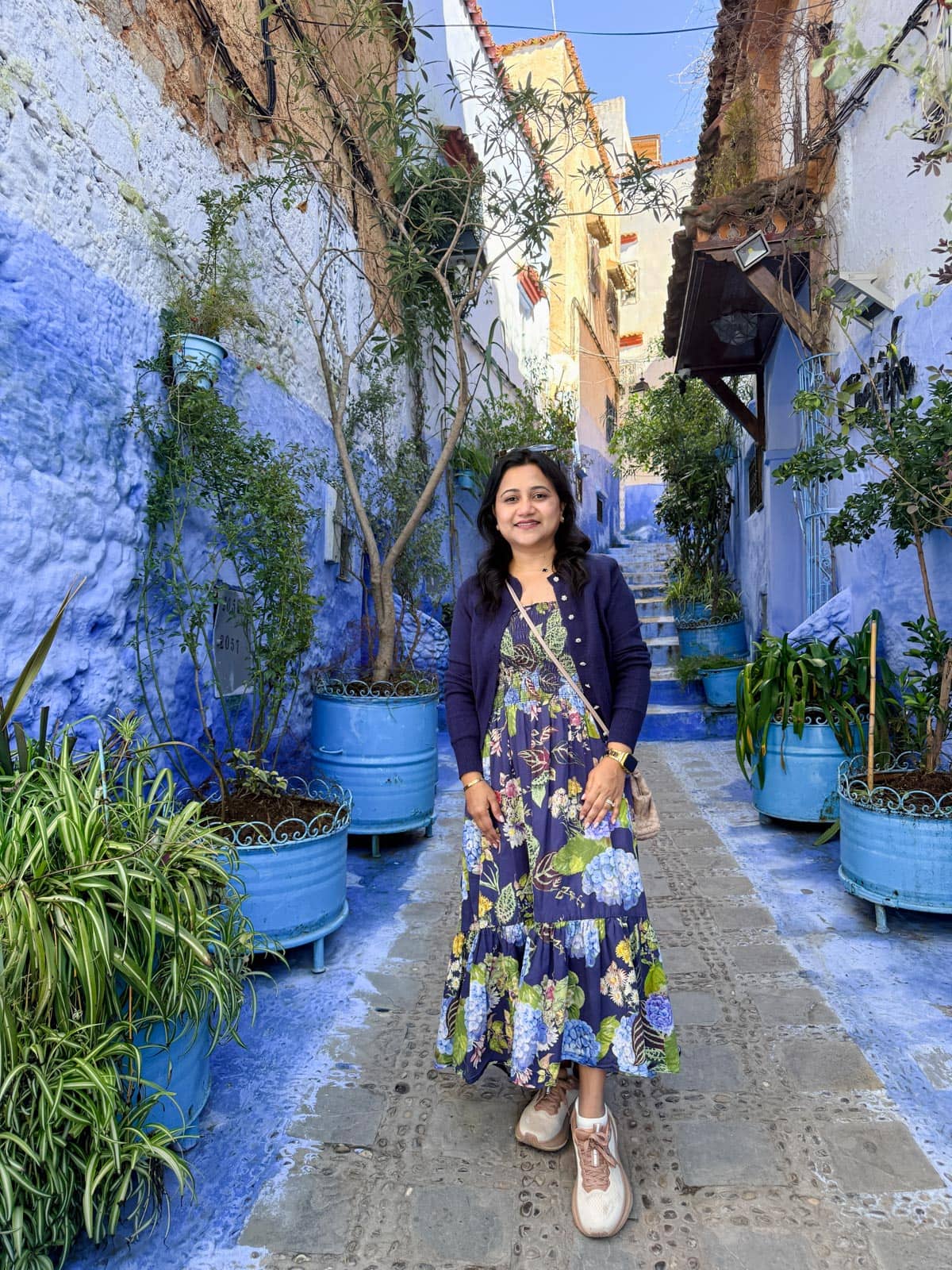 A woman in a floral dress stands on a narrow street lined with blue walls and blue potted plants in a sunlit, outdoor setting.