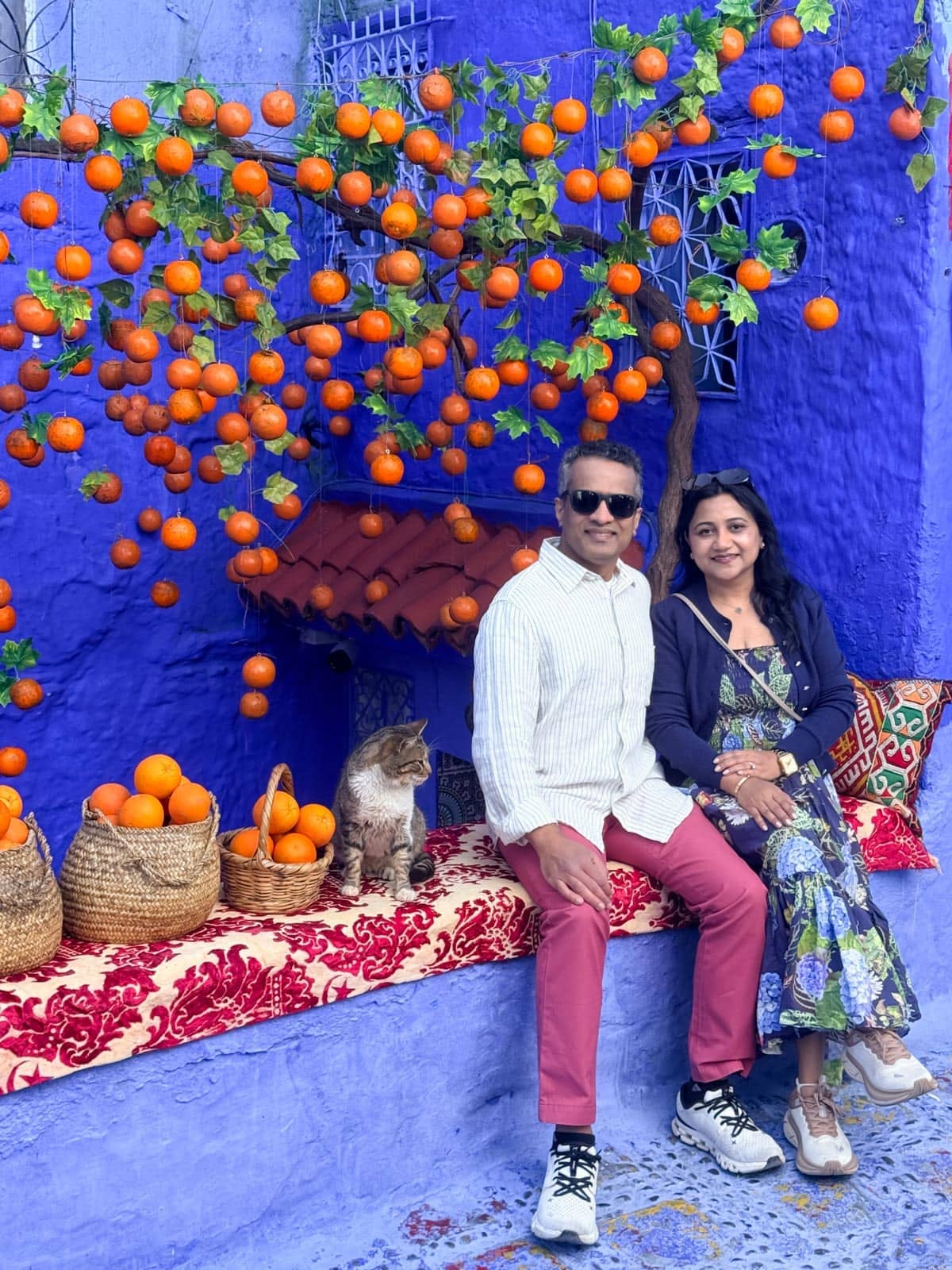 A man and woman sit on a red patterned bench by a purple wall with hanging oranges, baskets of oranges, and a cat beside them.