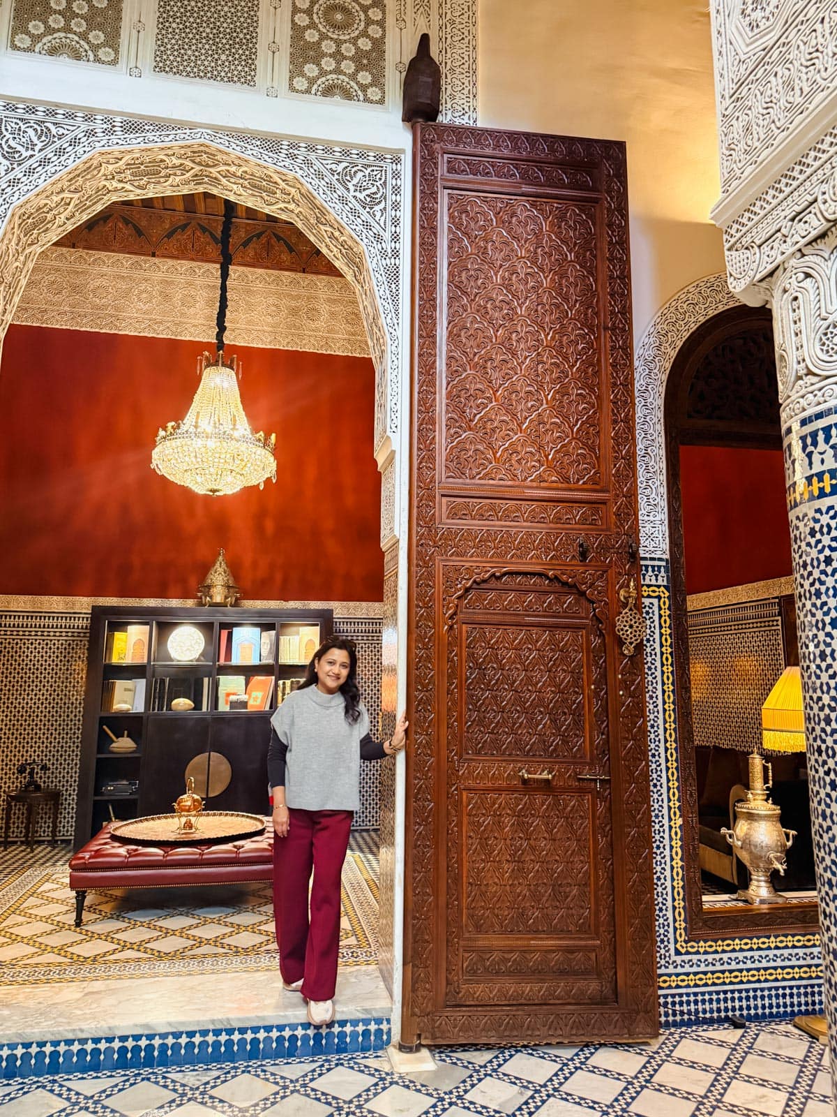 A woman stands next to a large, intricately carved wooden door in an ornately decorated room with mosaic tiles, arches, and a chandelier.