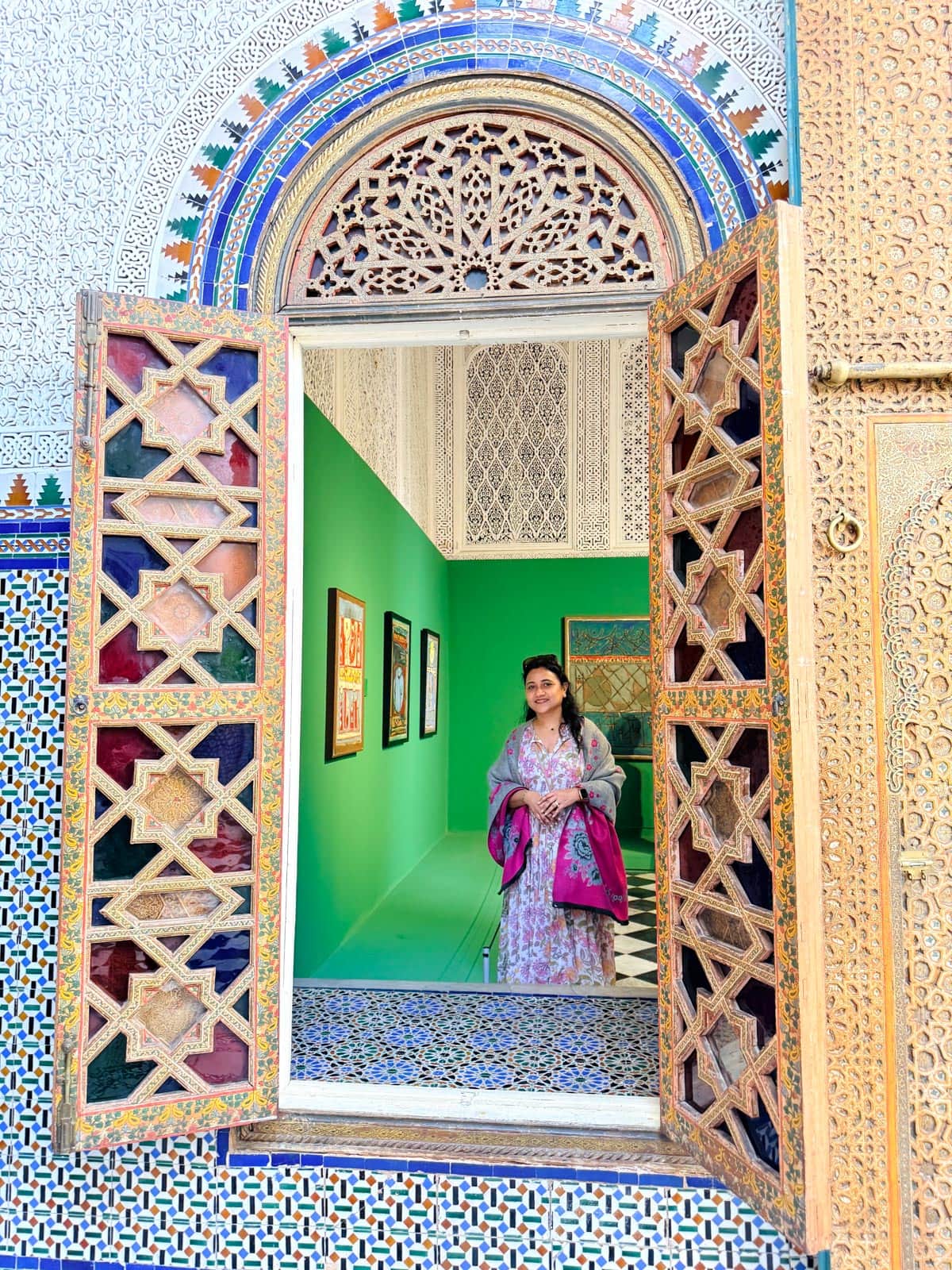 A woman in a patterned dress stands inside a colorful building, framed by an open ornate window with intricate geometric designs and mosaic tiles.