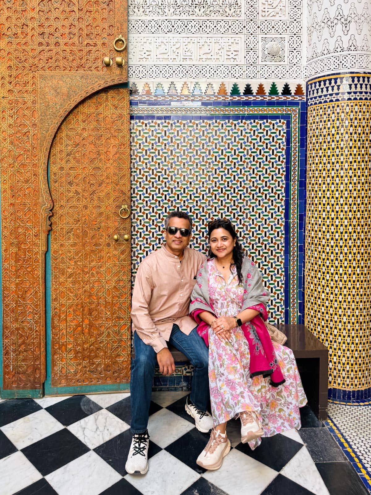 A man and a woman sit on a bench against an ornately tiled wall and carved wooden door, both smiling at the camera. The floor has a black and white checkered pattern.