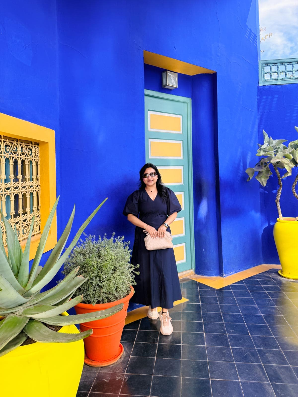 A woman in a dark blue dress stands in front of a vibrant blue wall with a green and yellow door, surrounded by large potted plants.