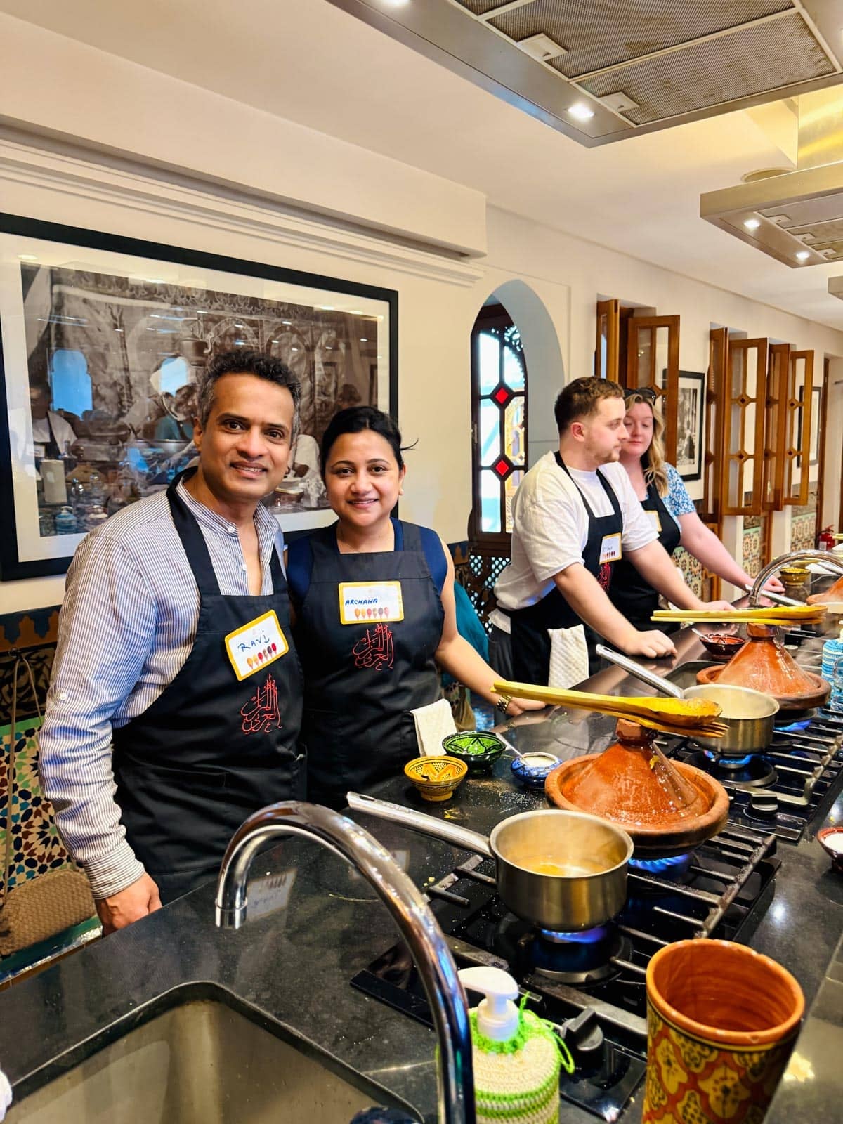 Four people in a kitchen, wearing aprons and name tags, stand behind a counter with pots and cooking utensils, appearing to participate in a cooking class.