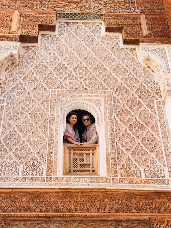 Two people stand at a carved window of an ornate, intricately patterned building with detailed stonework.