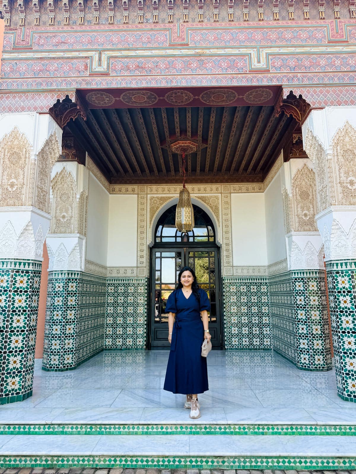 A woman in a navy dress stands on tiled steps in front of an ornate building with intricate green and white mosaic tiles and carved arches.