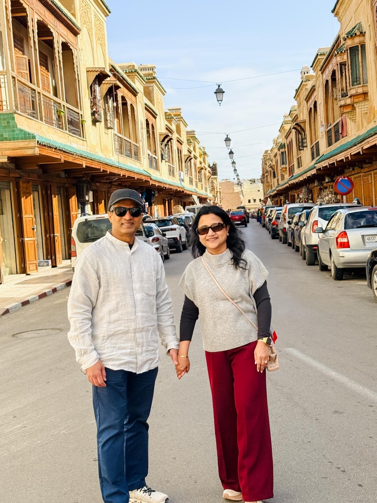 A man and woman stand holding hands on a street lined with parked cars and traditional wooden buildings under a clear sky.