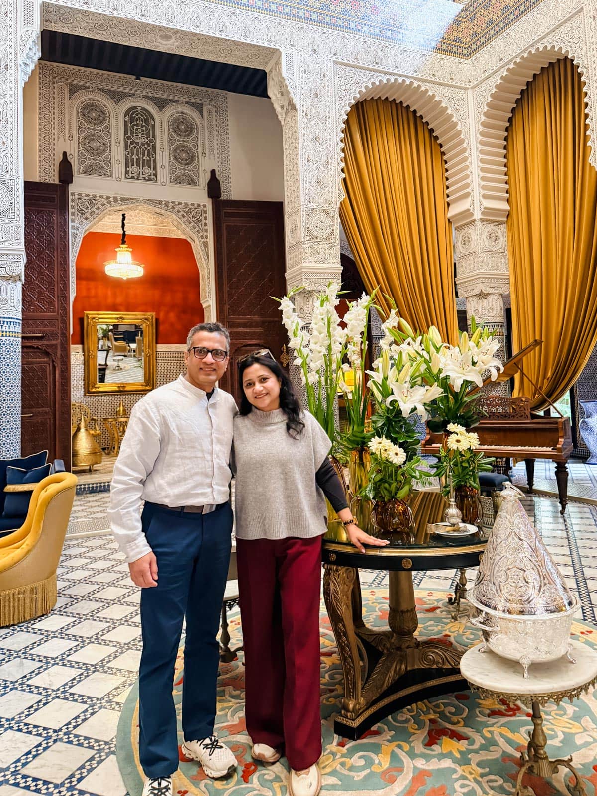 A man and woman stand together beside a round table with flowers in an ornately decorated room with patterned walls, arched doorways, and gold curtains.