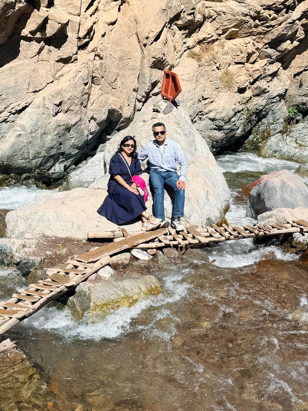 Two people sit on a large rock beside a flowing river, with a small wooden footbridge crossing the water in front of them and rocky cliffs in the background.