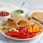 A plate with sliced donar kebab, grilled red and yellow bell peppers, onions, with flatbreads, a green sauce, and pickled vegetables on the side.
