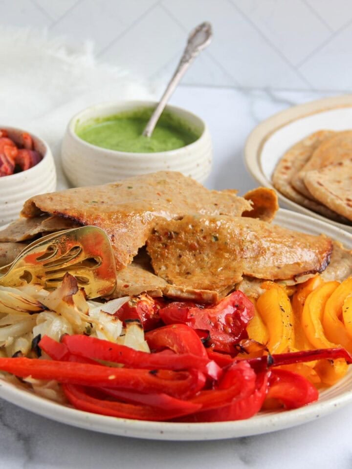 A plate with sliced donar kebab, grilled red and yellow bell peppers, onions, with flatbreads, a green sauce, and pickled vegetables on the side.