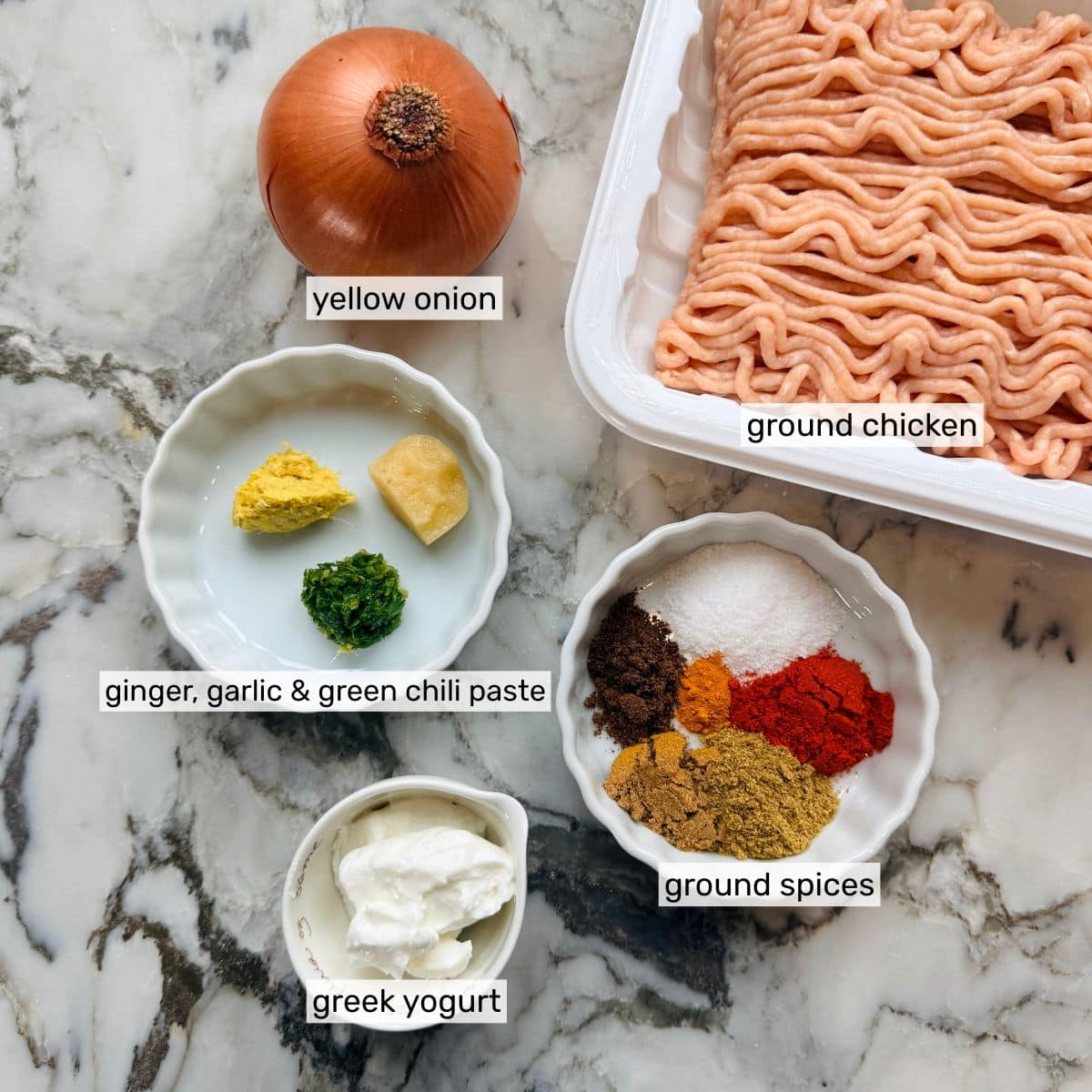 Ingredientes sobre una superficie de mármol: cebolla amarilla, pollo molido, pasta de jengibre, ajo y chile verde, yogur griego y un plato con diversas especias molidas.