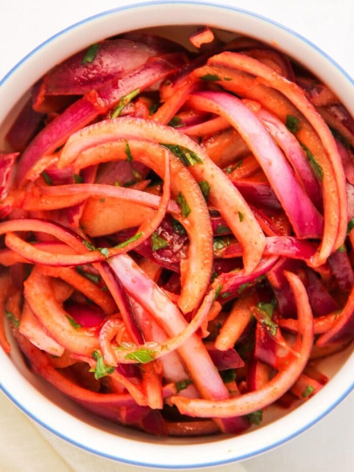 A white bowl filled with sliced red onions and herbs, mixed with a red seasoning or dressing.