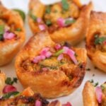 Close-up of golden-brown pastry puffs filled with chicken and topped with chopped herbs and pickled onions, labeled Bombay Chicken Burger Puffs.
