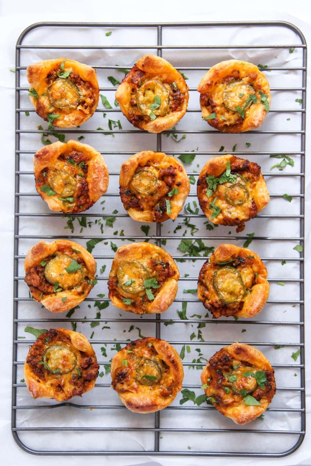 Twelve mini savory tarts with golden crusts, filled with melted cheese and herbs, arranged on a cooling rack over a white surface with chopped herbs scattered around.