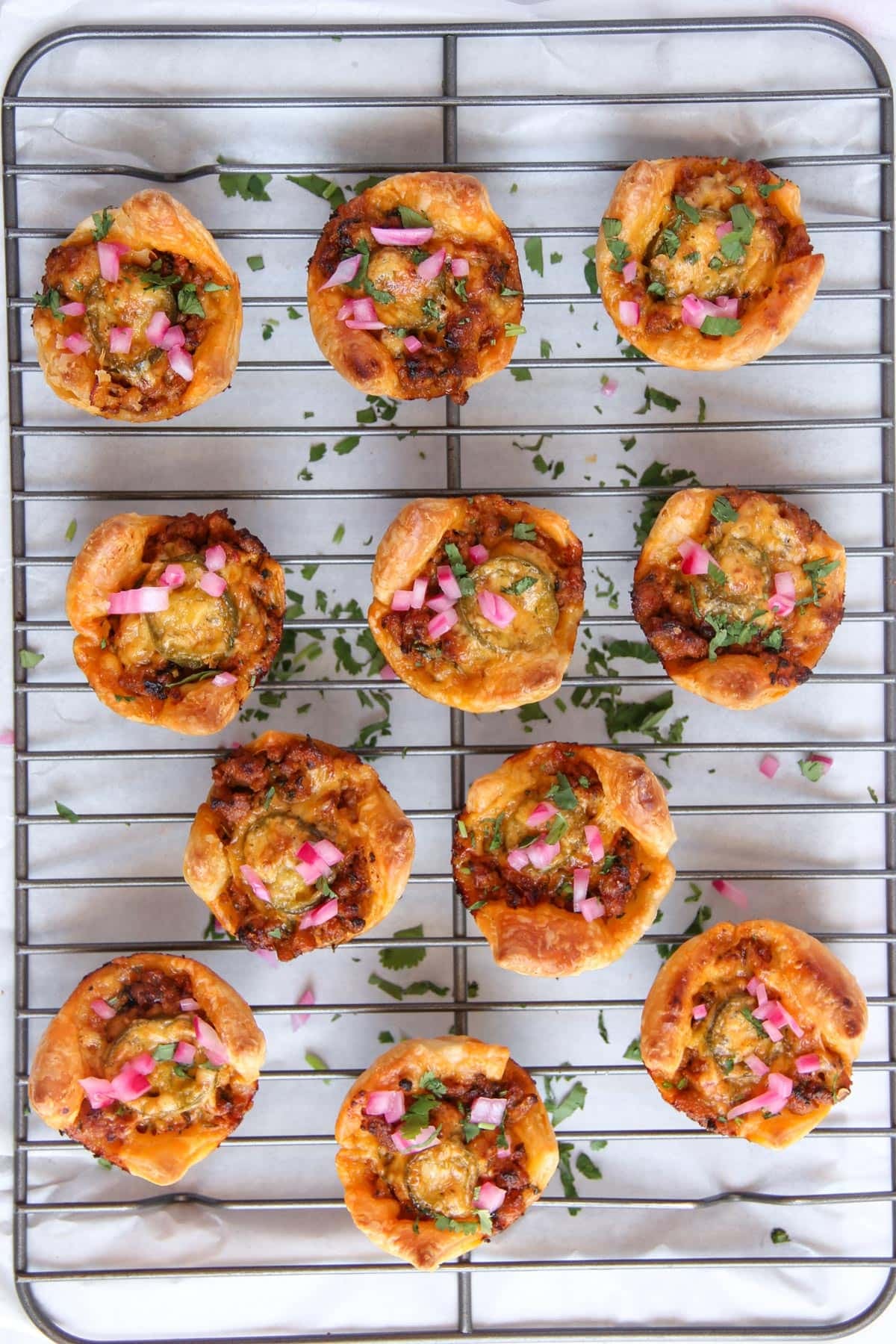 Twelve mini savory tarts topped with herbs and diced pickled onions are arranged on a metal cooling rack, with chopped herbs scattered over.