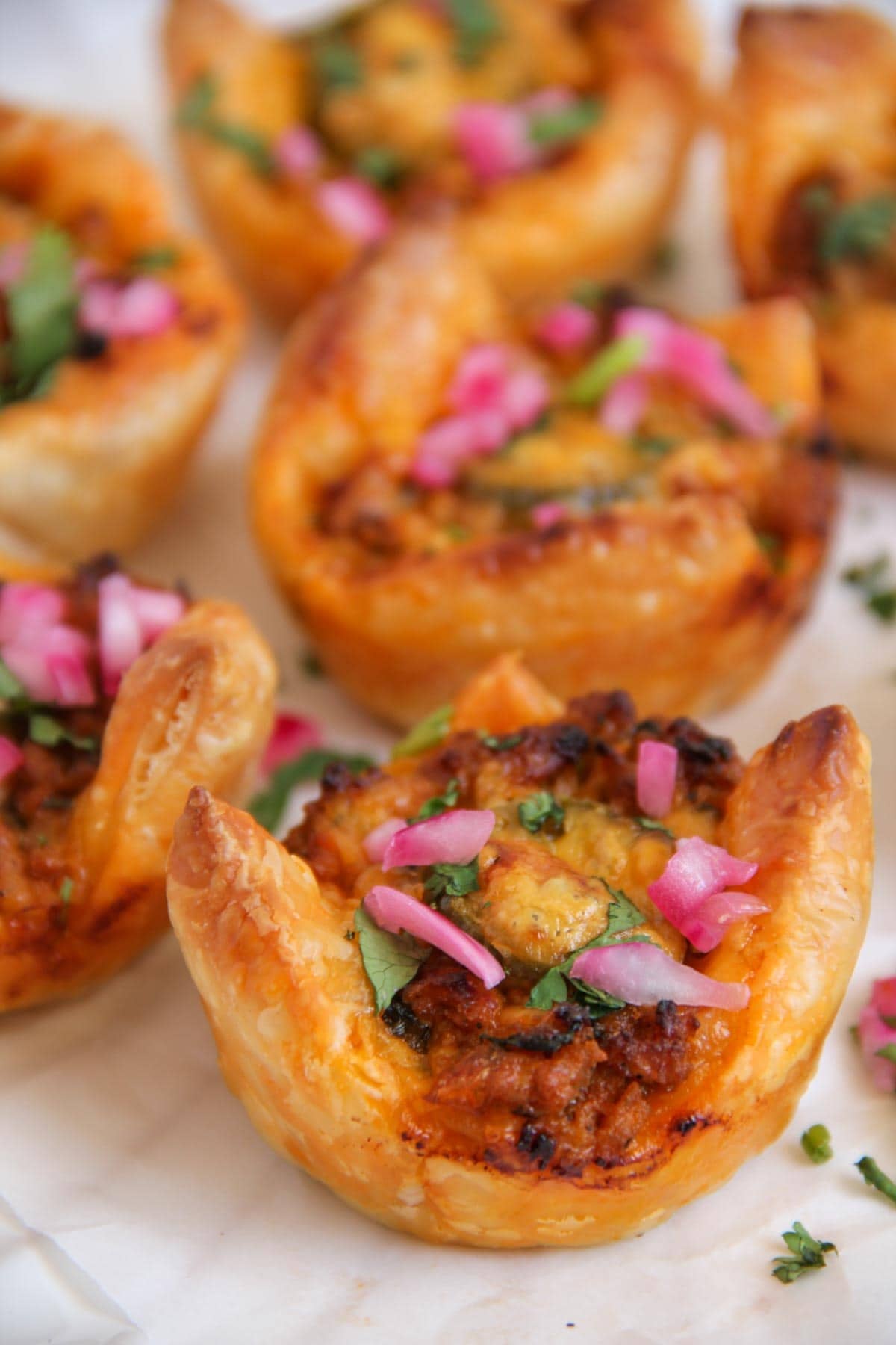 Close-up of baked puff pastry cups filled with a savory mixture, garnished with chopped herbs and pickled red onions, placed on parchment paper.