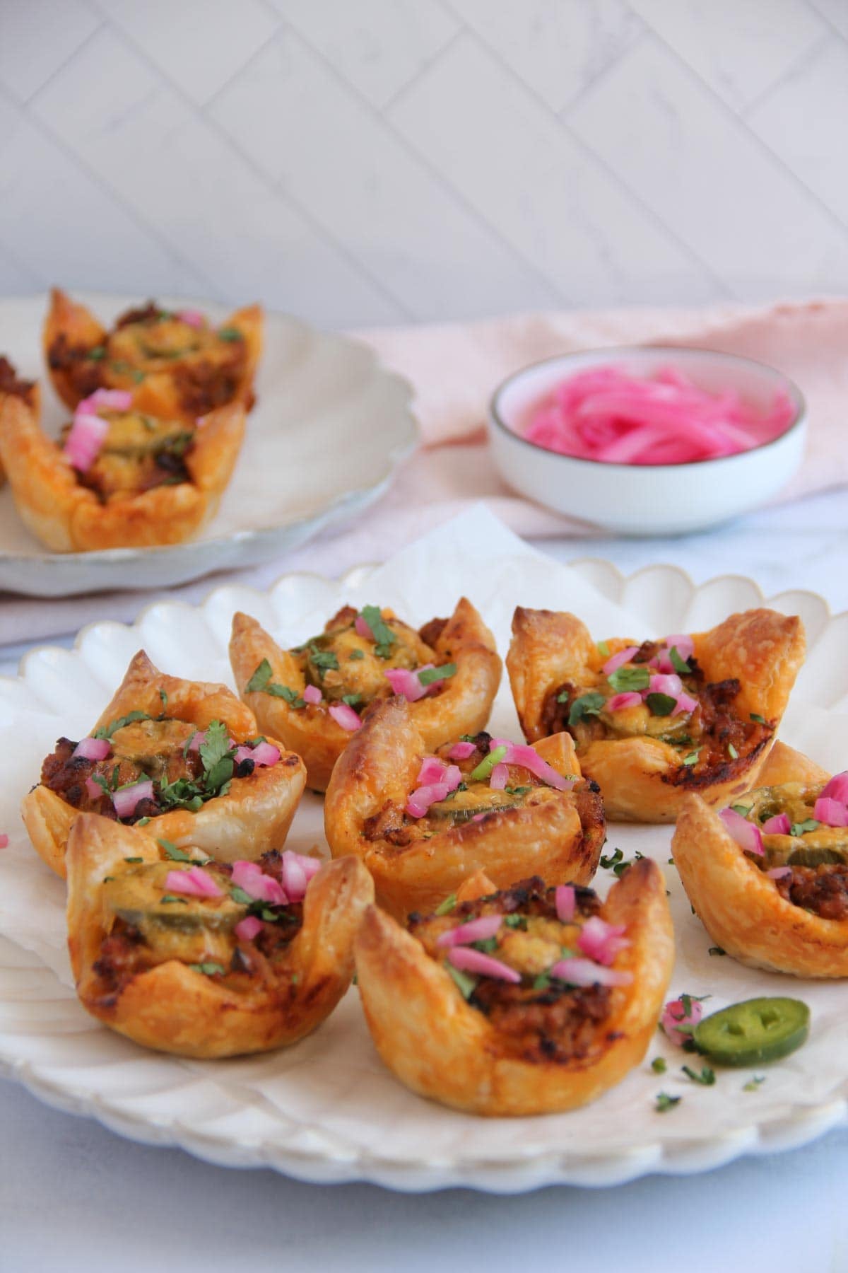 A plate of puff pastry cups filled with savory ingredients, garnished with chopped herbs and pickled onions, with a bowl of pickled onions in the background.