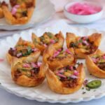 A plate of puff pastry cups filled with a savory mixture and garnished with chopped herbs and pickled onions, with more pastry cups and a bowl of pickled onions in the background.