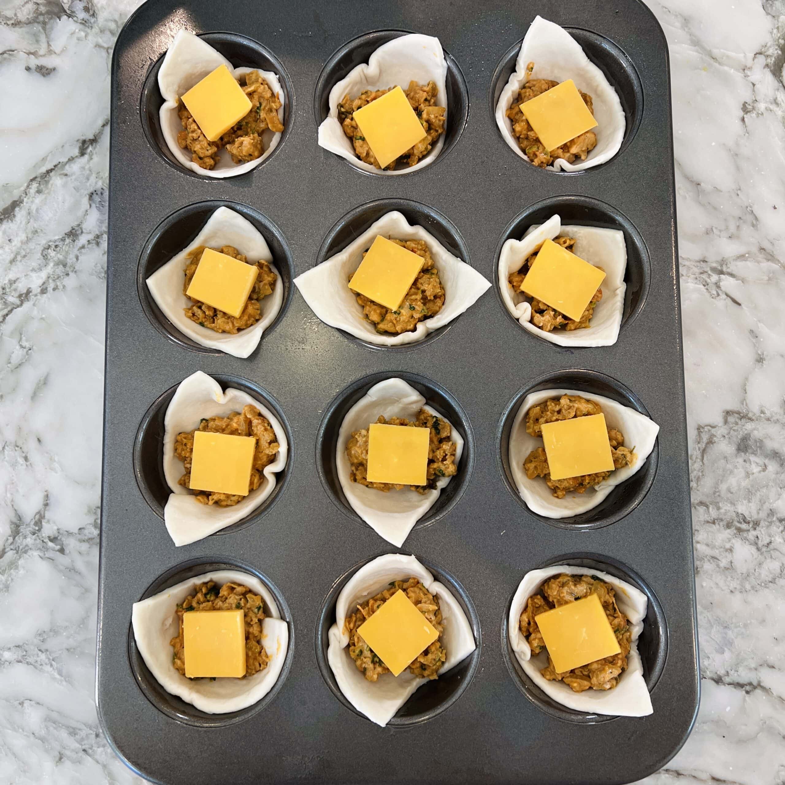 A muffin tin with twelve cups, each lined with pastry, filled with a meat mixture, and topped with a square slice of cheese, on a marble countertop.