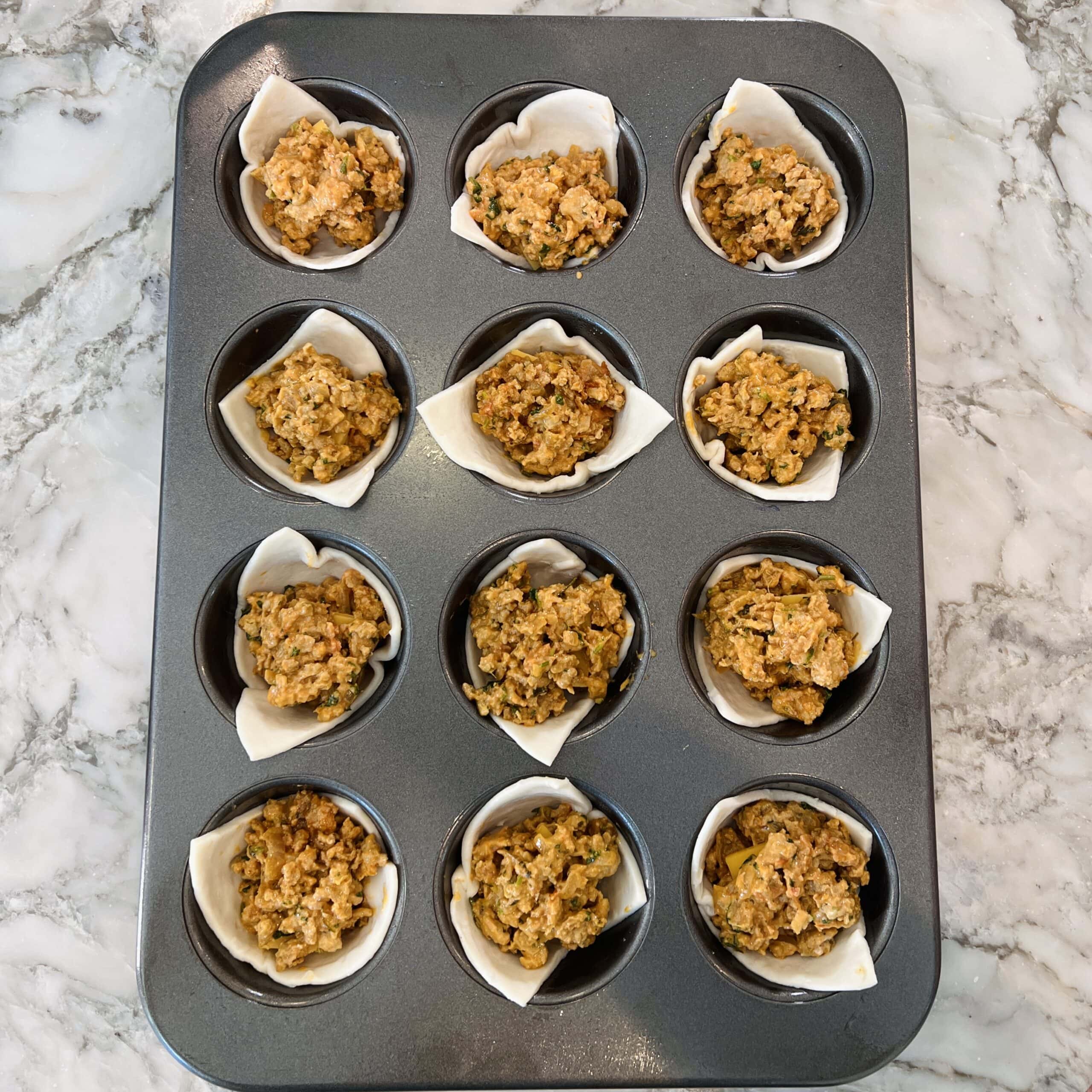 A muffin tin with 12 cups, each lined with pastry and filled with keema, sits on a marble countertop.