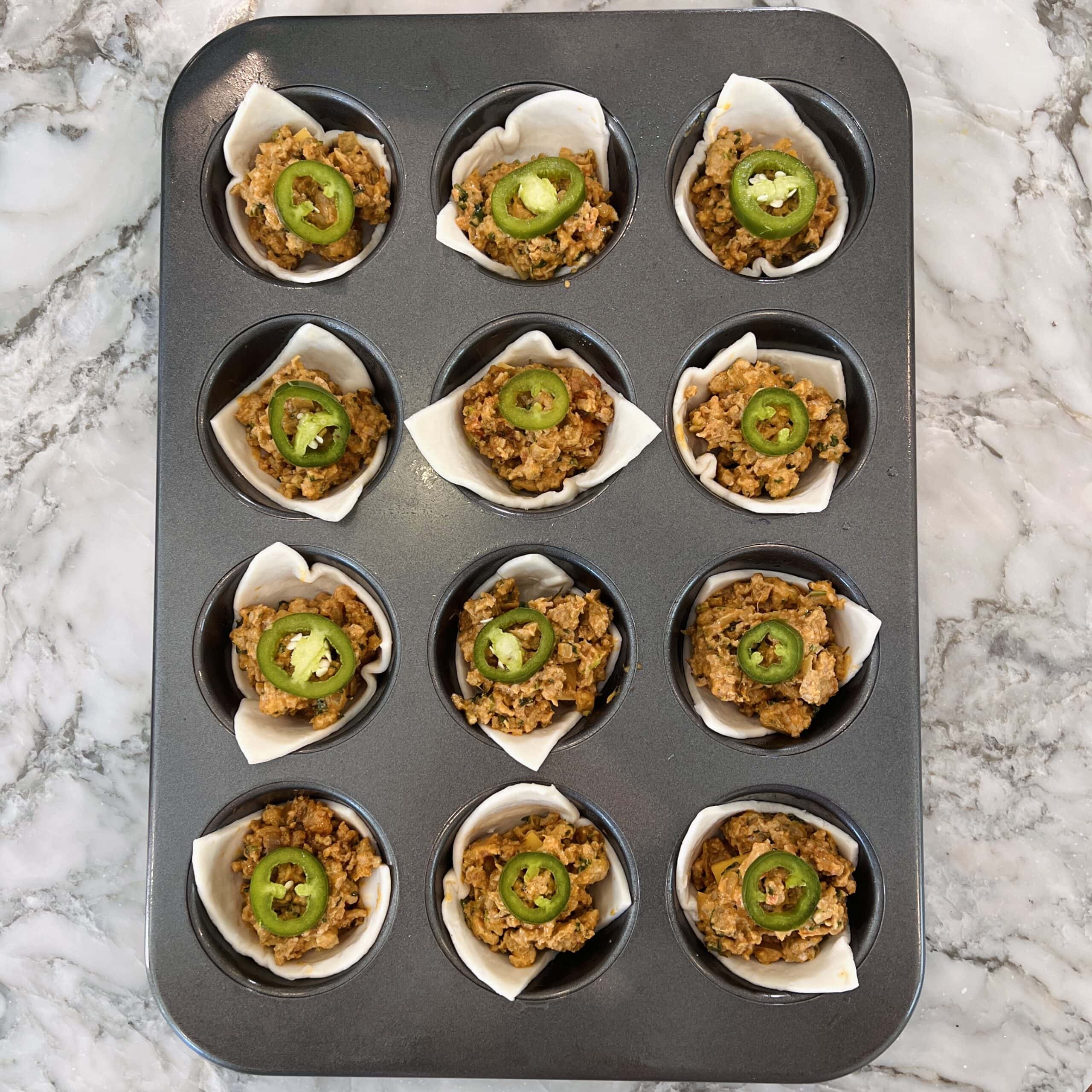 A muffin tin holds twelve paper-lined cups filled with a savory ground mixture, each topped with a slice of green jalapeño, on a marble countertop.