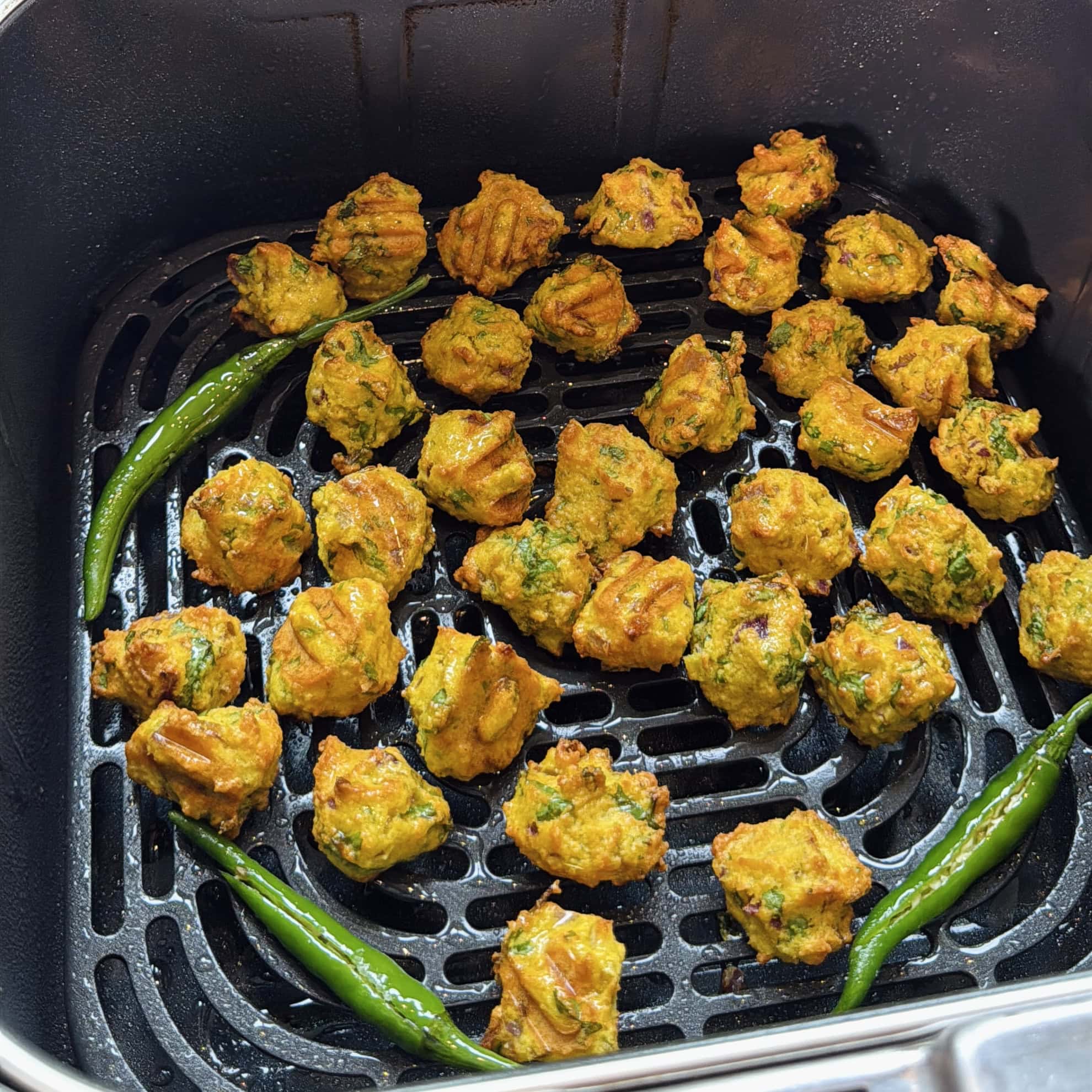 Golden brown fritters and whole green chilies arranged in an air fryer basket for cooking.