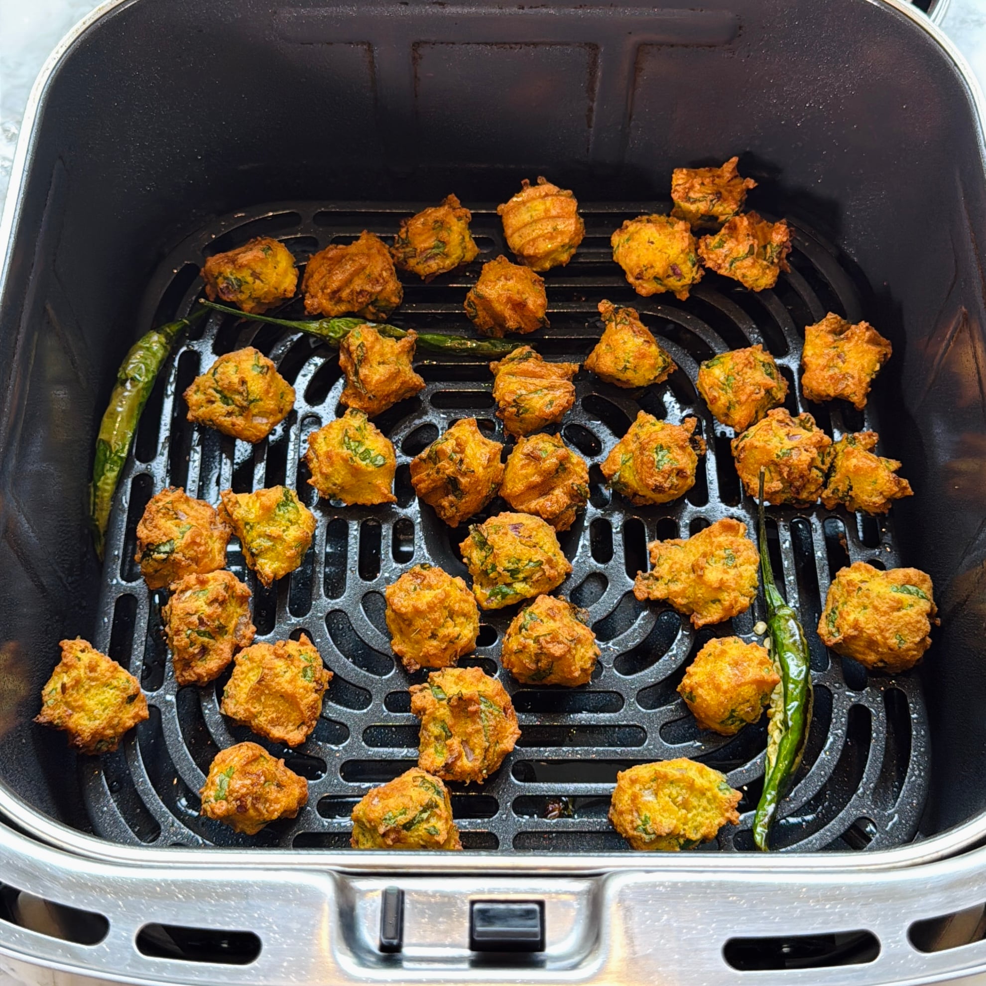 Golden brown vegetable fritters and whole green chilies are arranged in a single layer inside an air fryer basket.