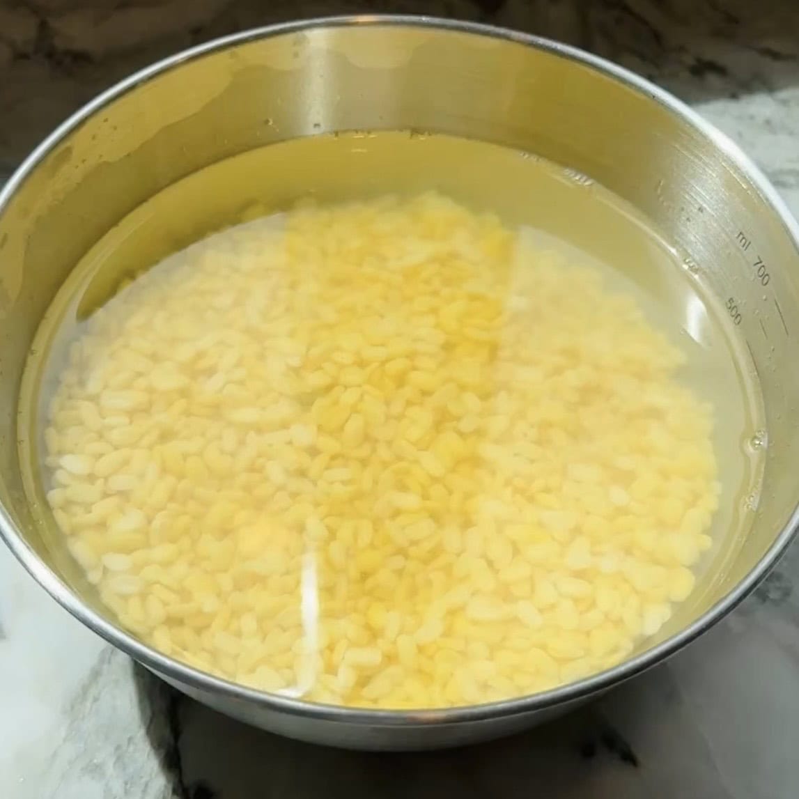 A stainless steel bowl filled with yellow split lentils soaking in water on a marble surface.