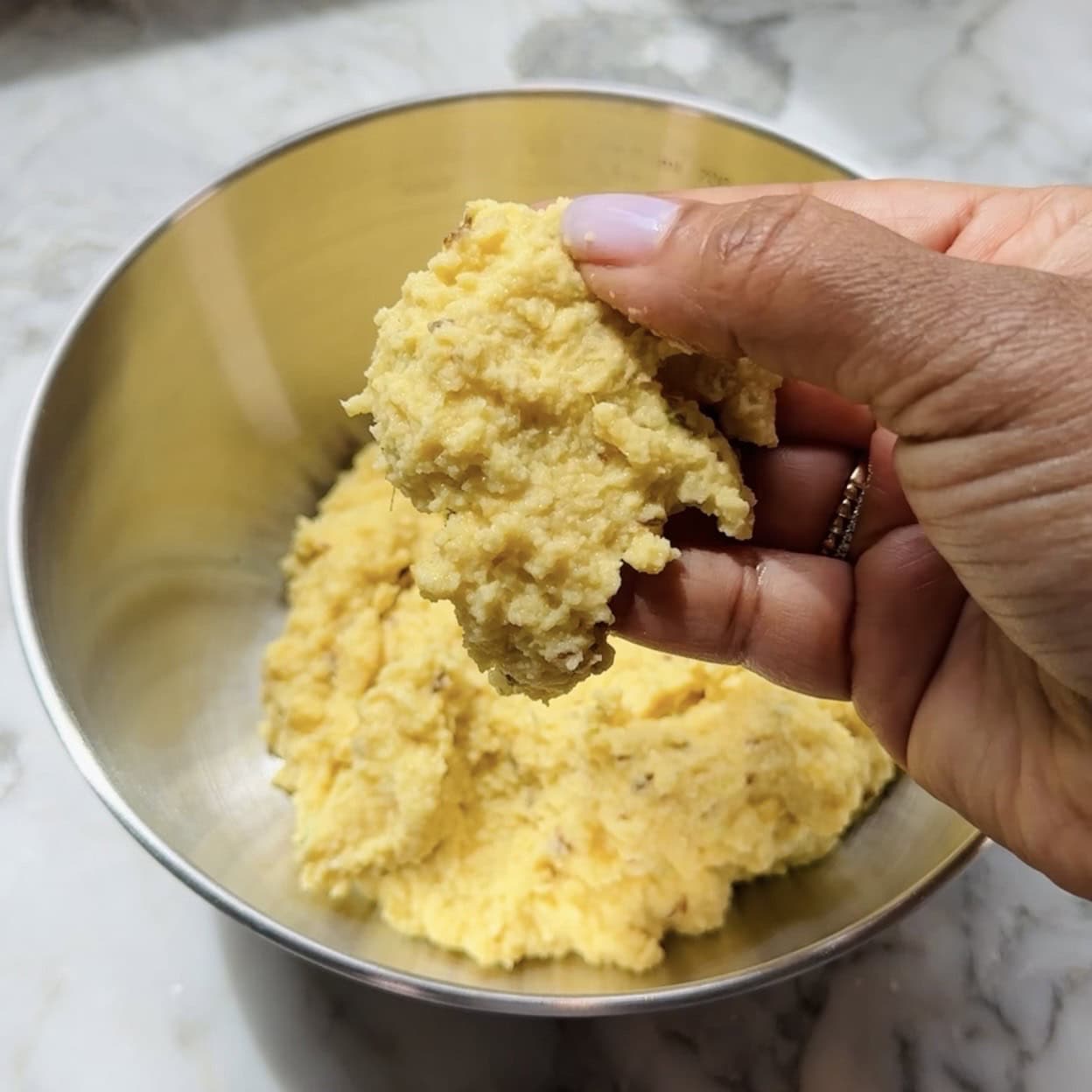 A hand holds a portion of yellow dough above a metal mixing bowl containing more dough, on a marble countertop.