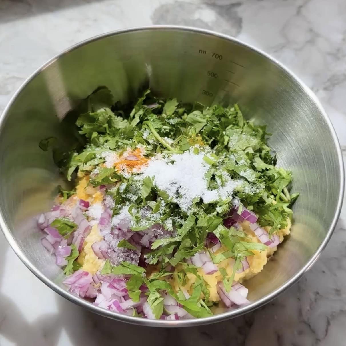 A stainless steel bowl containing chopped red onions, cilantro, spices, and salt on top of blended mumg dal.