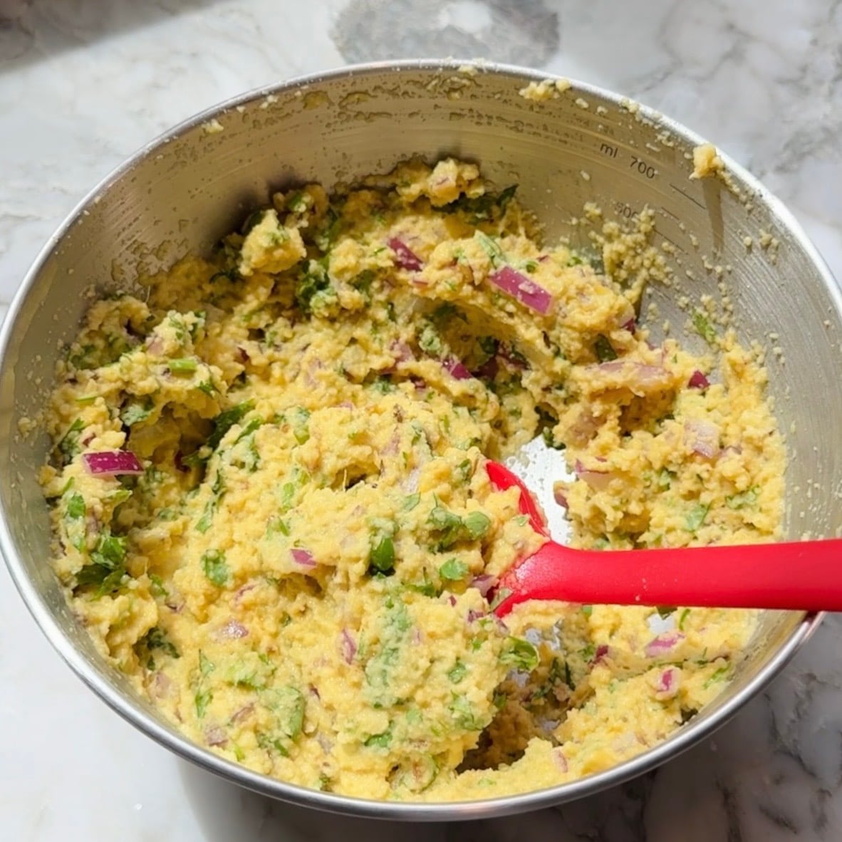 A metal bowl filled with a coarse, yellow batter mixed with chopped red onions and green herbs. A red spatula rests inside the bowl on a marble countertop.