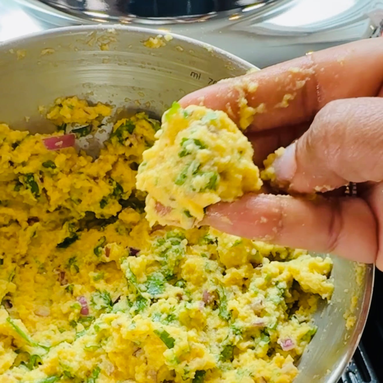 A hand holds a portion of a yellow, herb-filled dough mixture over a bowl containing more of the same mixture.